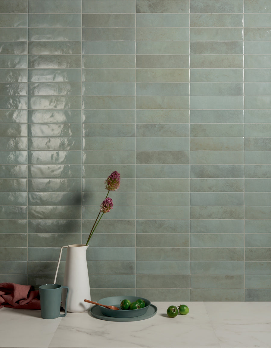 A minimalist kitchen scene with a green tile backsplash, a white pitcher holding two pink flowers, a blue cup, a plate with green limes, and a wooden spoon on a white marble countertop.