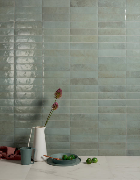 A minimalist kitchen scene with a green tile backsplash, a white pitcher holding two pink flowers, a blue cup, a plate with green limes, and a wooden spoon on a white marble countertop.