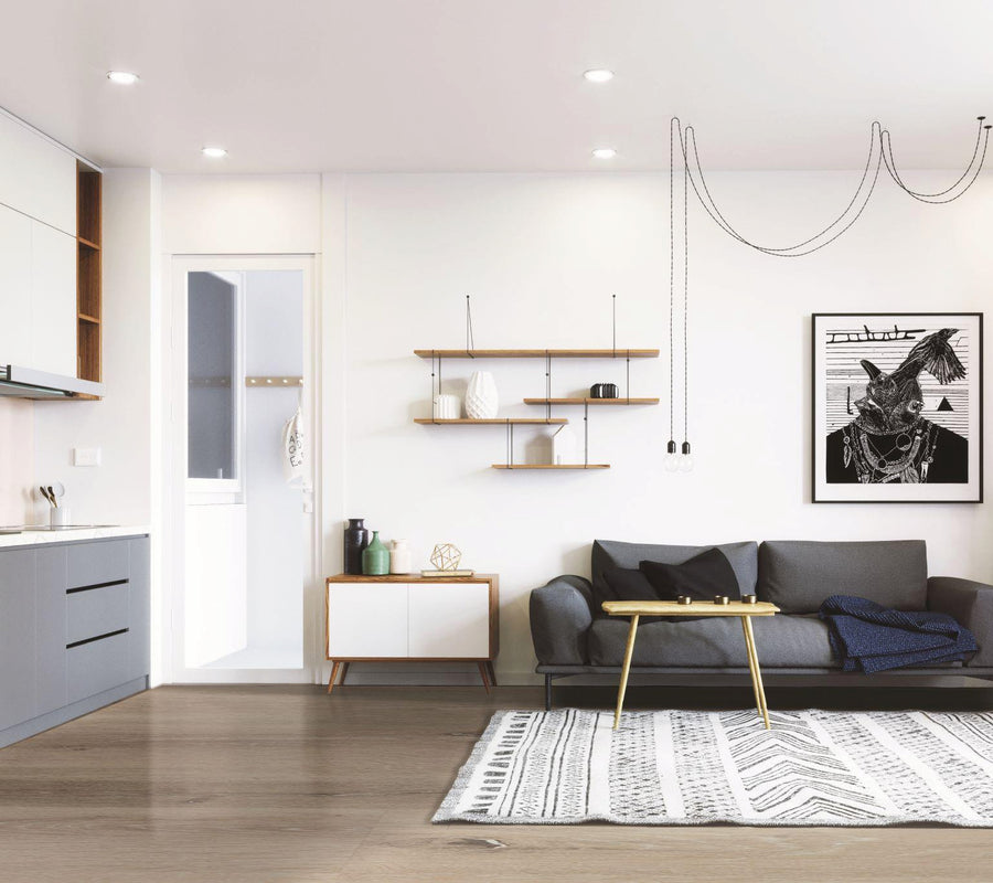 Modern living room with a gray sofa, geometric rug, wall art, floating shelves, and a small cabinet. Minimalist décor, with a kitchen area to the left and hanging pendant lights from the ceiling.