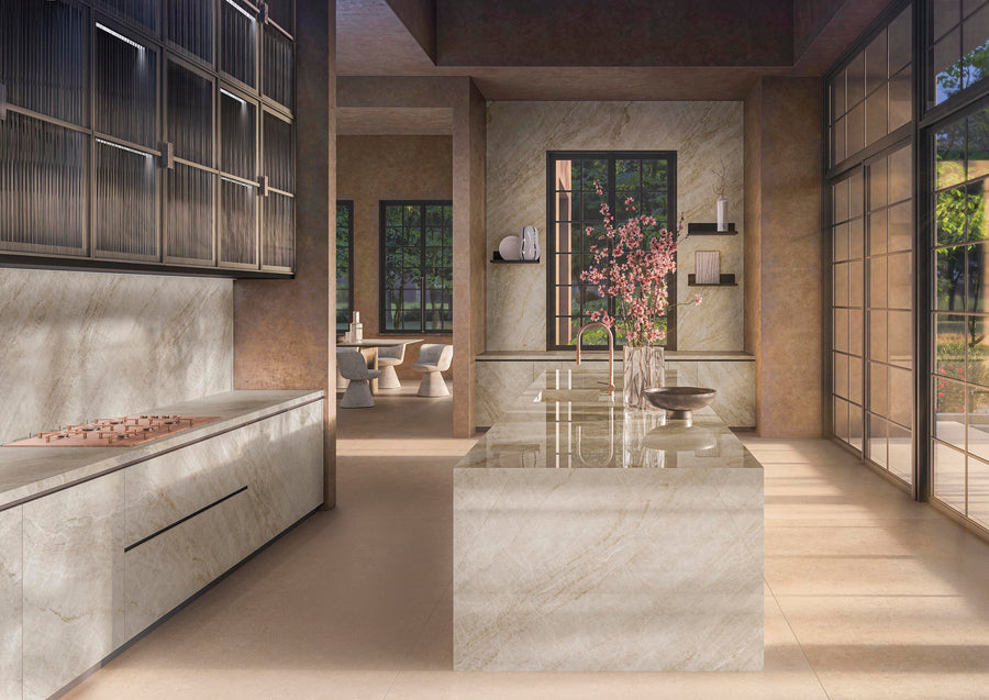 Modern kitchen with sleek, light marble surfaces, a large island with flowers in a vase, minimalist cabinets, and large windows letting in natural light. Dining area with round table and chairs visible in the background.