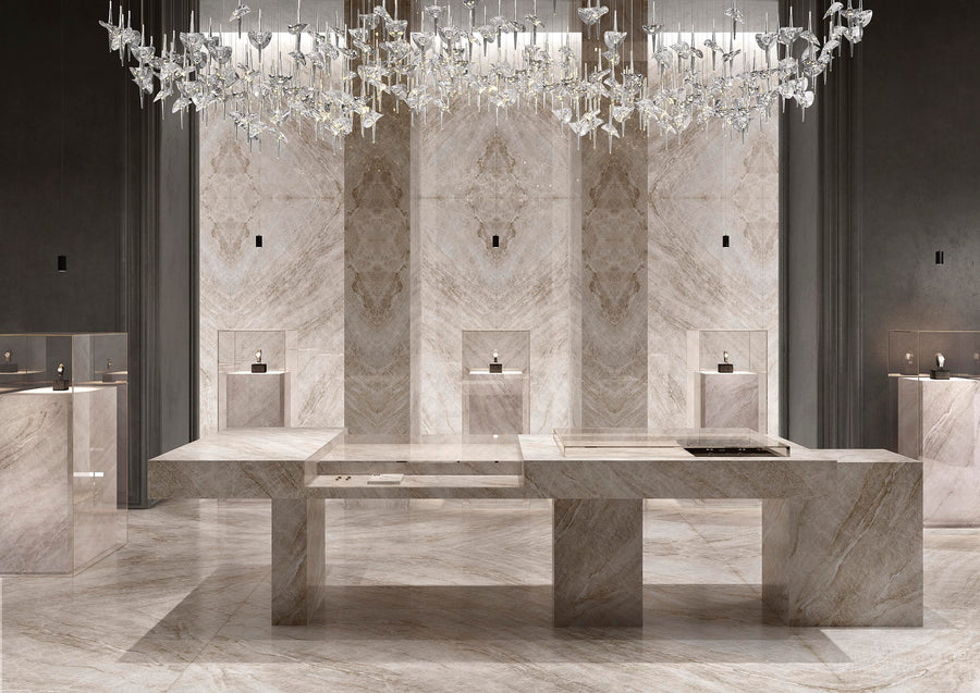 Luxurious showroom with polished marble floors, counters, and walls in beige tones, a modern geometric table, small display cases, and an elegant crystal chandelier overhead.