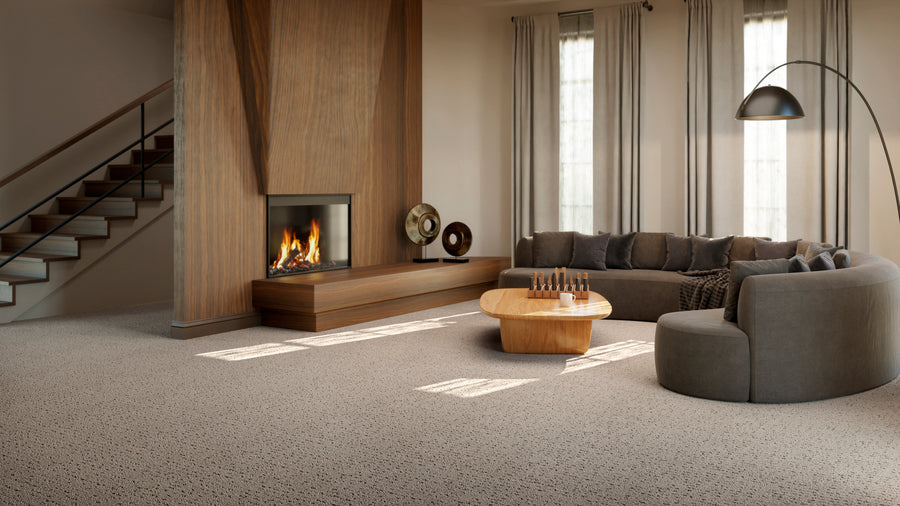 A modern living room with a curved gray sofa, wooden coffee table, fireplace, abstract decor, and large windows with light curtains. Carpeted floor and a staircase are visible on the left.