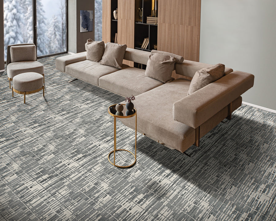 Modern living room with a beige sectional sofa, matching ottoman, round gold side table, textured gray patterned carpet, wooden shelving unit, and large window showing snowy trees outside.