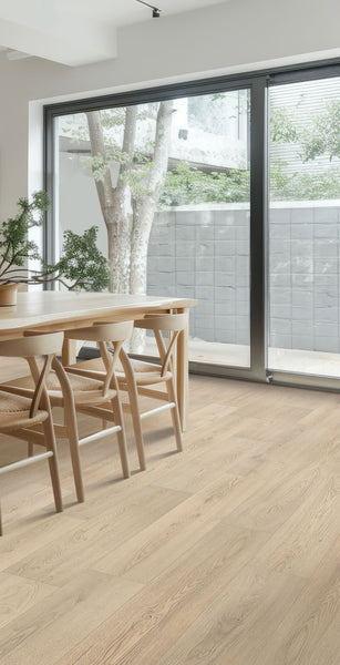 A minimalist dining area with light wooden chairs and table, potted plant centerpiece, large floor-to-ceiling windows, and an outdoor view of a tree and gray privacy wall.