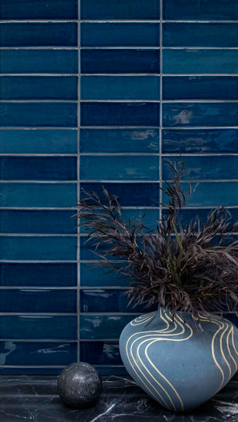 A blue vase with wavy yellow lines holds dark dried foliage, sitting on a black marble surface. A round, dark stone is nearby. The background features glossy, rectangular blue tiles.