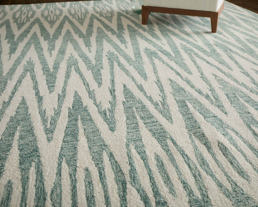 A close-up of a rug with a white and light blue zigzag pattern. The corner of a wooden-legged chair with a white cushion is visible at the top right of the image.