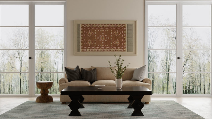 A living room with a beige sofa, black coffee table, wooden side table, and patterned rug. Large windows reveal trees outside. A decorative rug is framed and hung on the wall above the sofa.