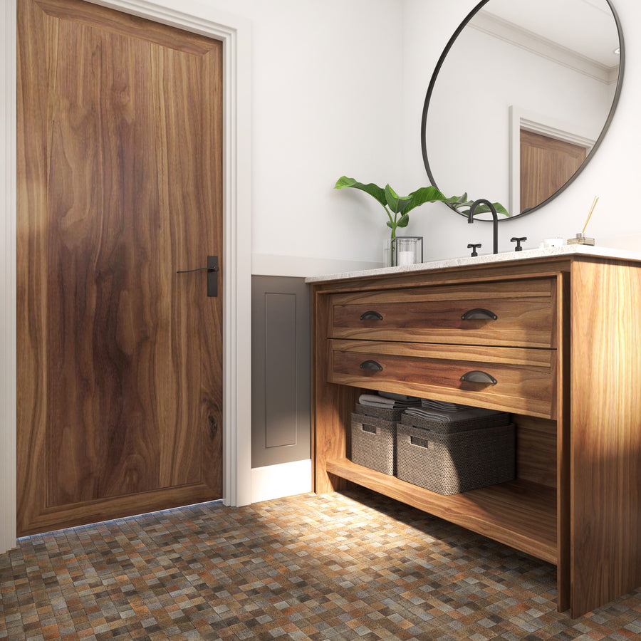 A modern bathroom features a wooden vanity with drawers, two woven baskets on the lower shelf, a round mirror, a black faucet, a plant in a vase, and a wooden door. The floor is covered with mosaic tiles.