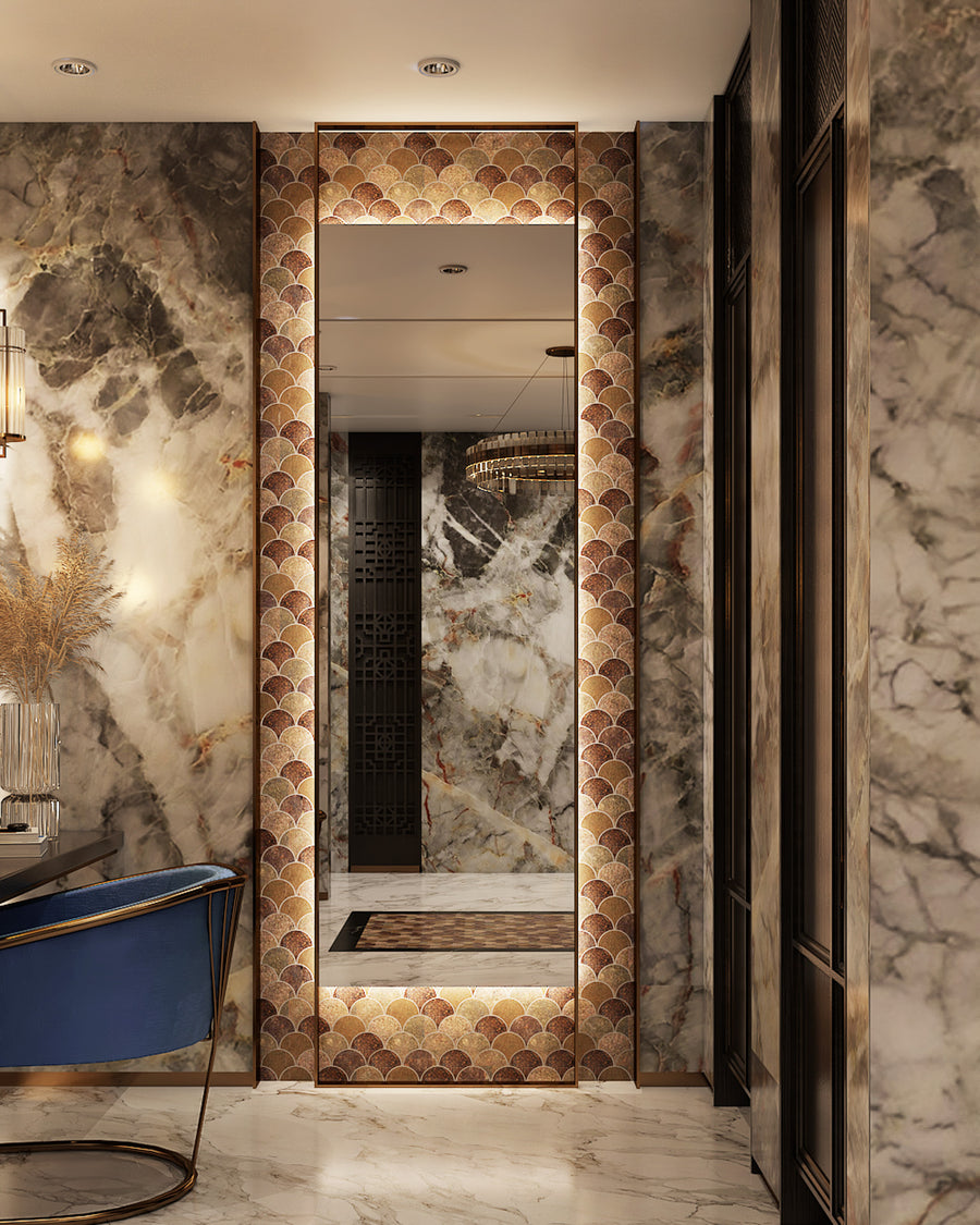 A luxurious hallway with marble walls and floor features a large, ornately framed mirror with a gold and bronze scallop pattern, reflecting a chandelier and another marble wall. A blue velvet chair is partially visible on the left.