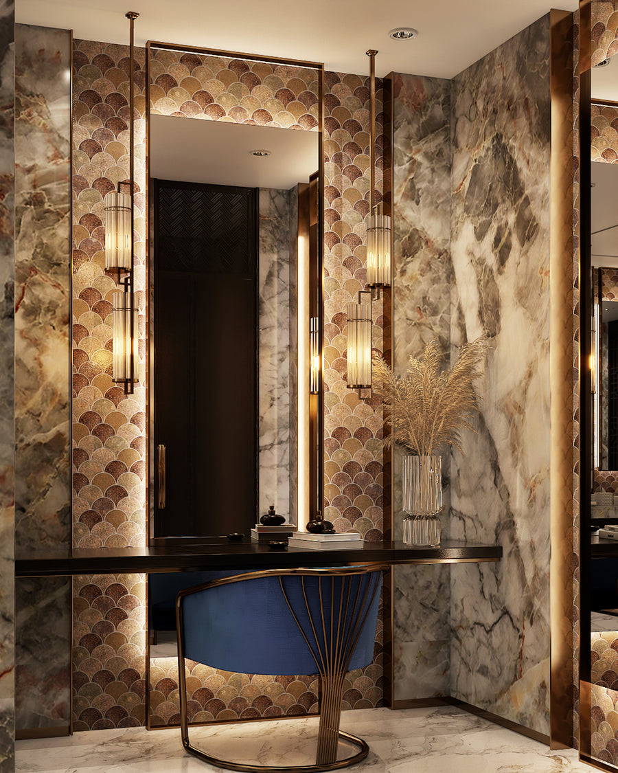 A luxurious vanity area with a marble wall, gold accents, patterned wallpaper, a dark desk, blue velvet chair, tall glass vases with dried grass, and warm vertical lighting fixtures.