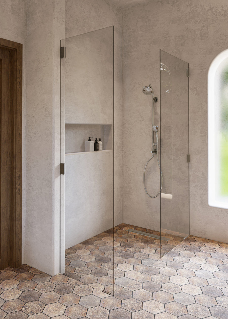 A minimalist bathroom with a walk-in shower featuring clear glass panels, a wall-mounted showerhead, and built-in shelving holding two bottles. The floor has hexagonal tiles, and sunlight streams through an arched window.