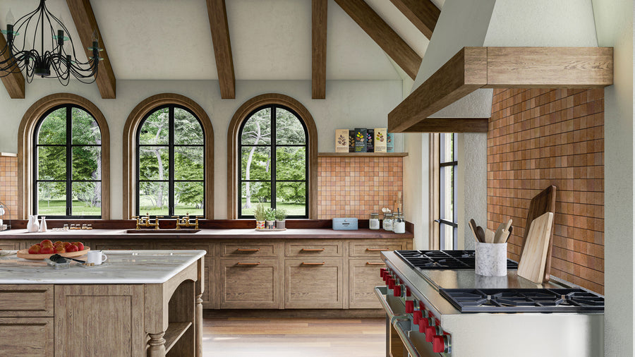 A rustic kitchen with wooden cabinets, exposed ceiling beams, arched windows with black frames, and a marble island. There are kitchen appliances and utensils on the countertops, with greenery visible outside.