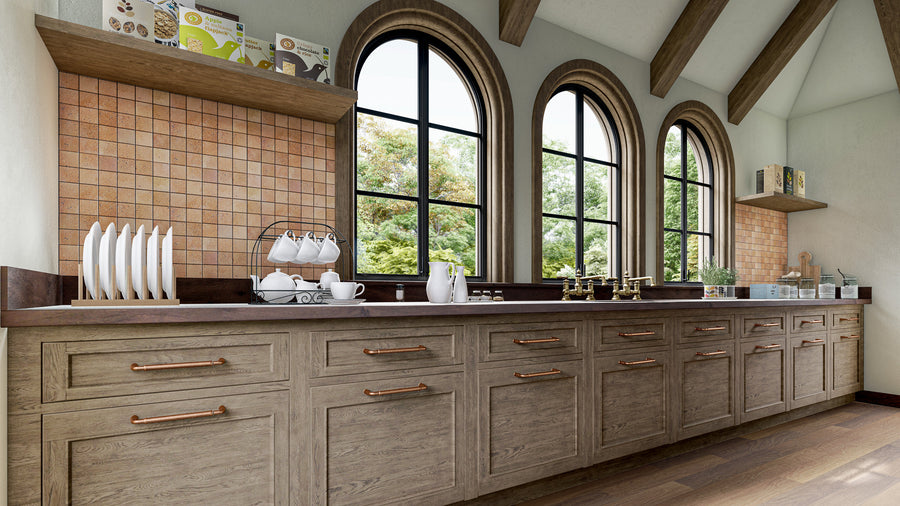A spacious kitchen with arched windows, wooden cabinets, copper handles, and a countertop with dishes, a tea set, and kitchen items. Sunlight streams in, highlighting the warm, natural tones of the room.