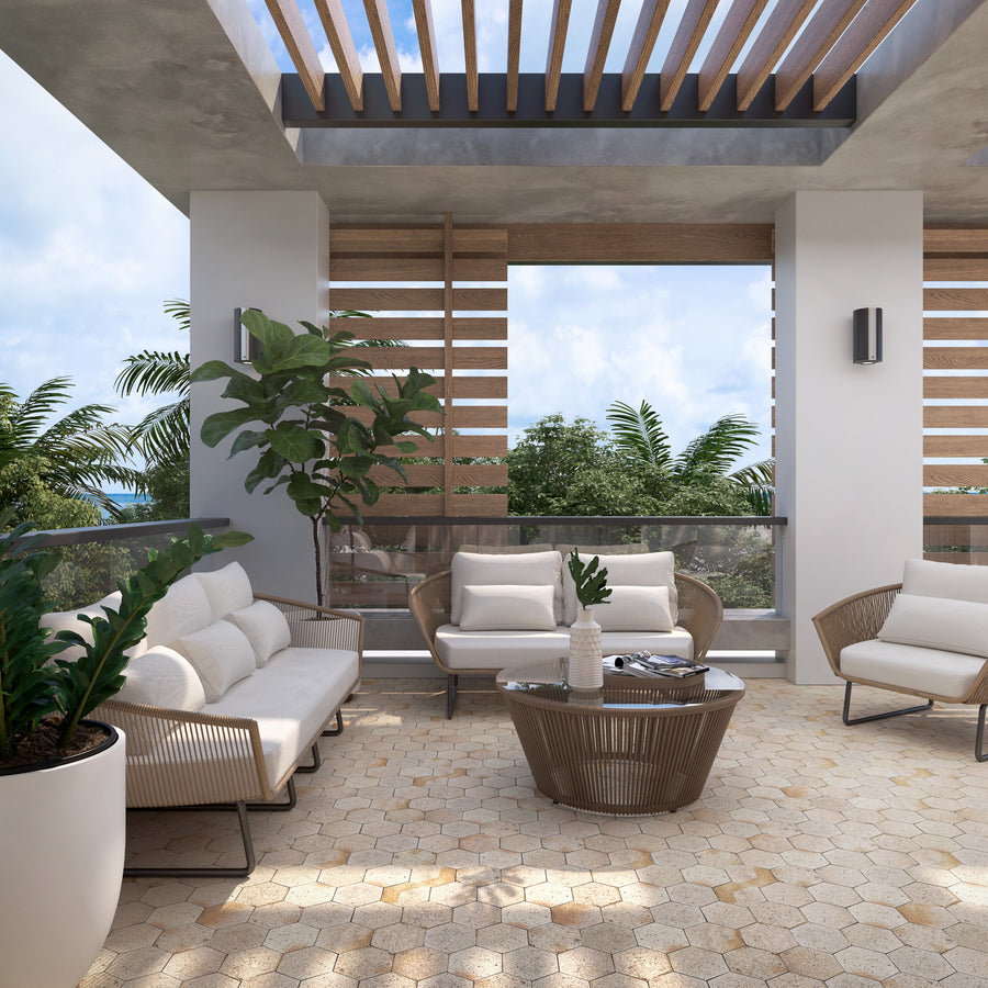 Modern outdoor patio with white cushioned seating, a round coffee table, potted plants, and wooden slat roof and walls. Sunlight filters through, and lush greenery is visible outside.