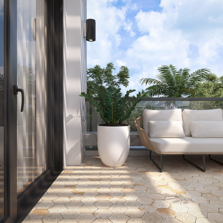 A modern balcony with hexagonal tile flooring, a potted plant, a cushioned outdoor sofa, glass railing, and greenery visible outside under a partly cloudy sky.