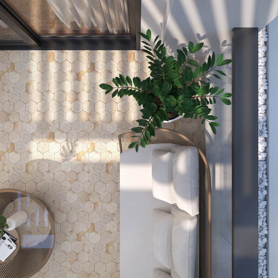 A top-down view of a cozy outdoor patio with hexagonal beige tiles, a white cushioned sofa, a green potted plant, a round table, a glass wall, and decorative white stones along the edge. Sunlight casts soft shadows.