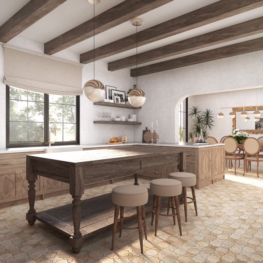 A rustic kitchen with exposed wooden beams, a large wooden island with three cushioned stools, beige cabinets, pendant lights, and a dining area with round table and chairs in the background. Sunlight streams through the window.