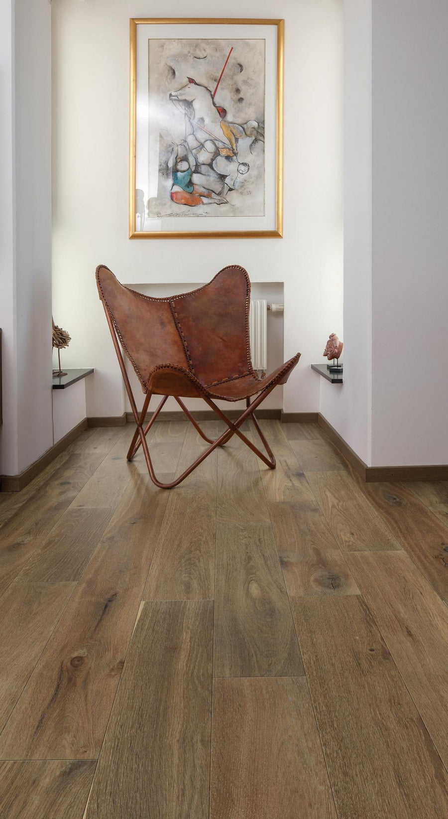 A brown leather butterfly chair sits on a wooden floor between two white walls. A framed abstract painting hangs on the wall above the chair. Small decorative objects rest on glass shelves flanking the chair.