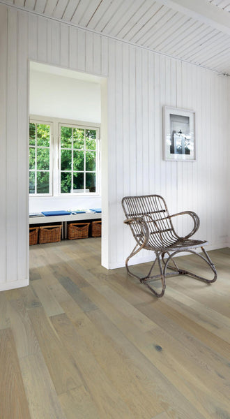 A bright, airy room with white paneled walls, light wood flooring, a wicker chair, and a large window with a view of green trees. Storage baskets sit beneath a bench by the window, and a framed picture hangs on the wall.