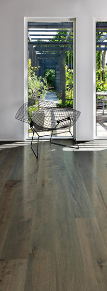A modern room with wide-plank wooden flooring, a minimalist black wire chair, and large windows opening to a lush garden with greenery and a pergola structure outside.