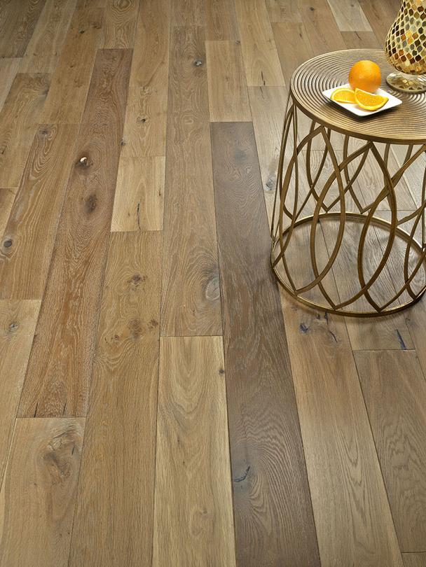 A round side table with a gold metal base and a glass top holding an orange and orange slices sits on a floor with light brown wooden planks.