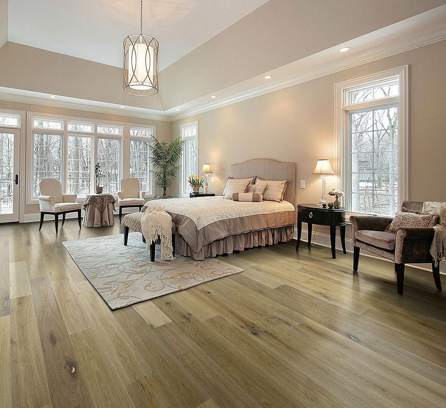 Spacious, elegant bedroom with large windows, light wood flooring, a king bed with beige bedding, two chairs, a plush rug, lamps, and decorative plants, all in neutral, warm tones.