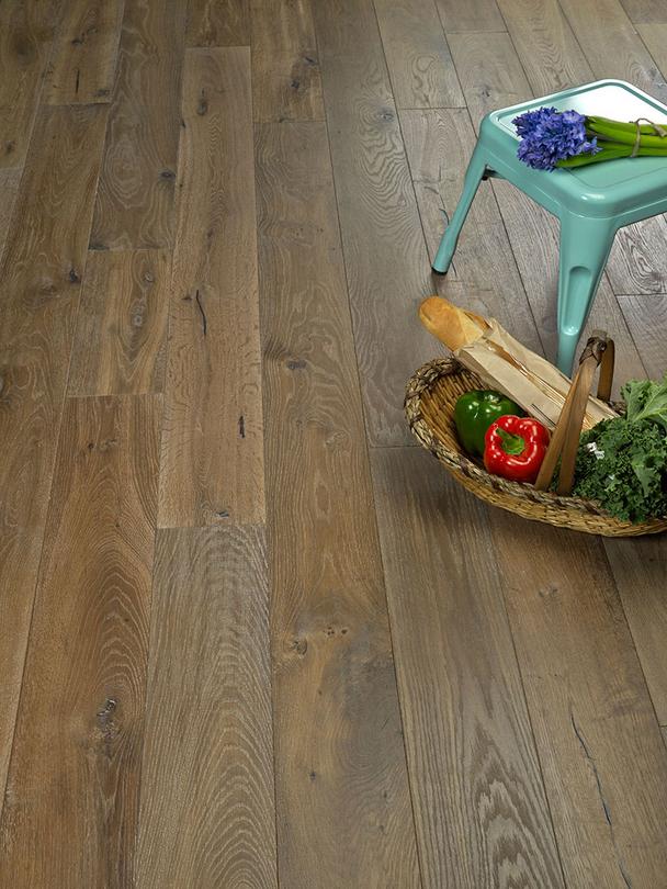 A woven basket with a baguette, kale, a red bell pepper, and a green pepper sits on wooden flooring next to a light blue metal stool holding purple flowers.