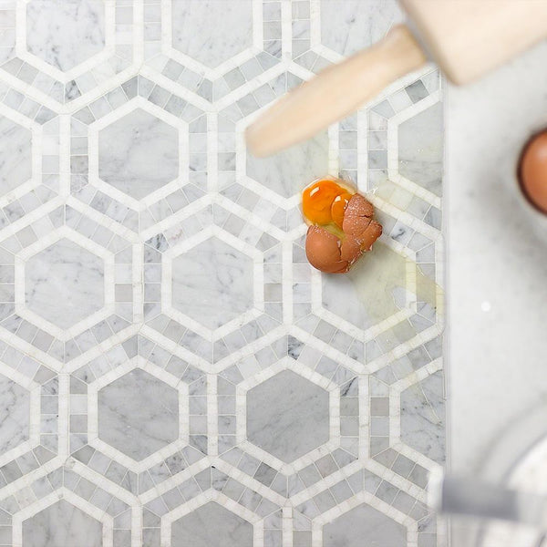 A cracked egg with spilled yolk on a white and gray geometric-patterned marble floor, next to a rolling pin and more eggs on a marble countertop.