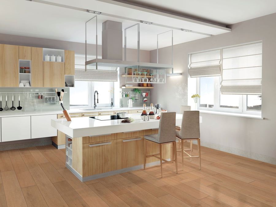 Modern kitchen with light wood cabinets, a large island with two beige chairs, built-in appliances, glass shelves with dishes, potted plants, and large windows with white blinds letting in natural light.