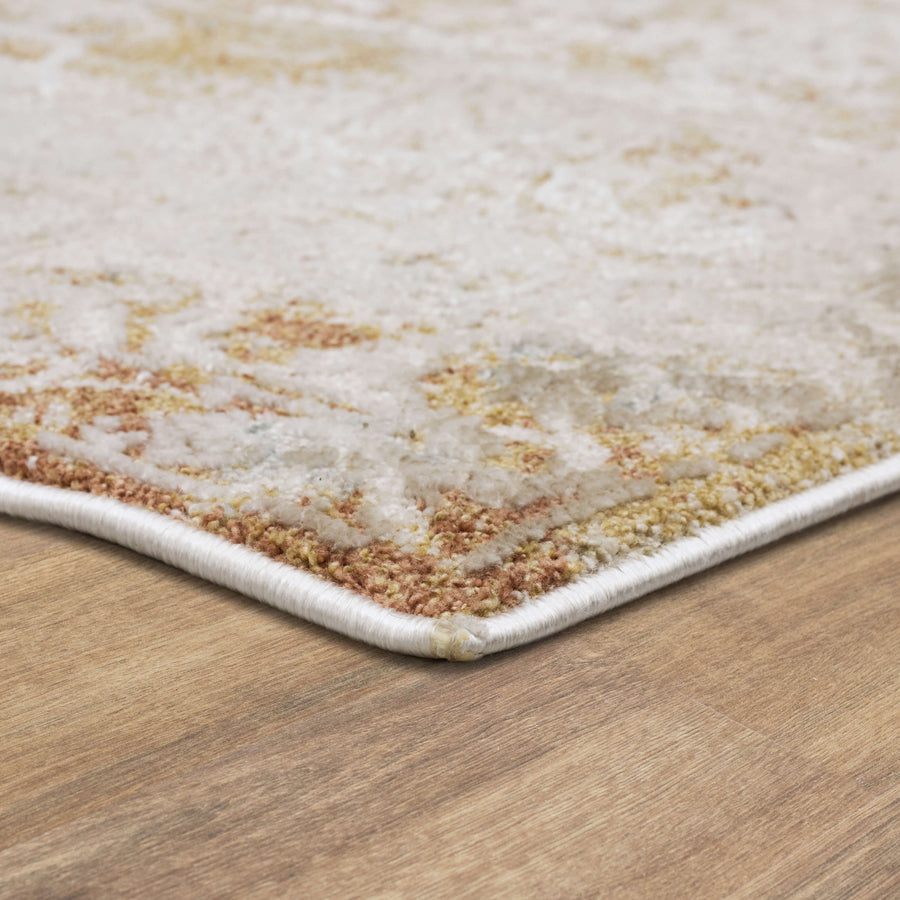 Close-up of the corner of a light-colored area rug with beige, cream, and golden tones, lying on a wooden floor. The rug edge is slightly rounded and neatly finished with white binding.