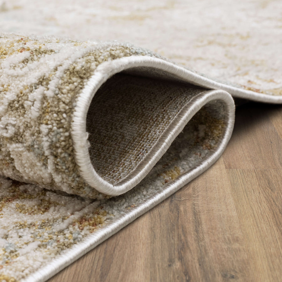 A close-up of a beige and light brown textured area rug with its corner folded over, showing the backing and soft pile, resting on a wooden floor.