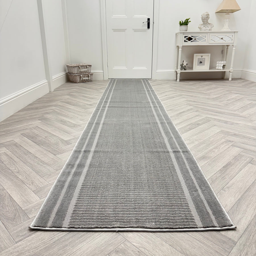 The Timeless 6931-900 Runner Rug, with subtle striped patterns and a gray hue, stretches along a light herringbone wood floor in a bright hallway, leading to a white door and accentuated by a wicker basket and white console table.