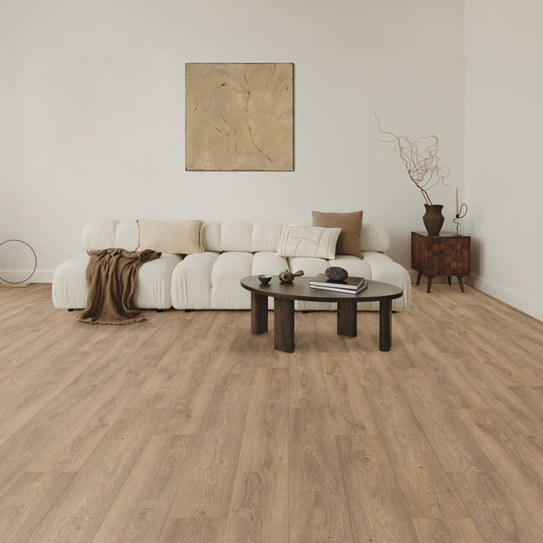 A modern living room with light wood flooring, a white sectional sofa with throw pillows and a brown blanket, a round dark coffee table, abstract wall art, and a small side table with decorative branches.