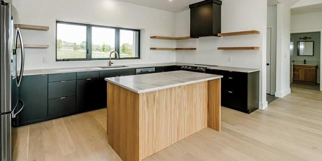 Modern kitchen with light wood floors, black cabinets, open wood shelves, a large island with a marble countertop, stainless steel appliances, and a window over the sink letting in natural light.