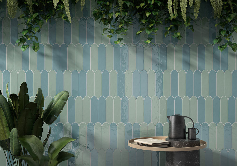 A modern interior with a wall of glossy, blue-green scalloped tiles, hanging greenery above, a large leafy plant on the left, and a round table with a black pitcher, cups, and books in the foreground.