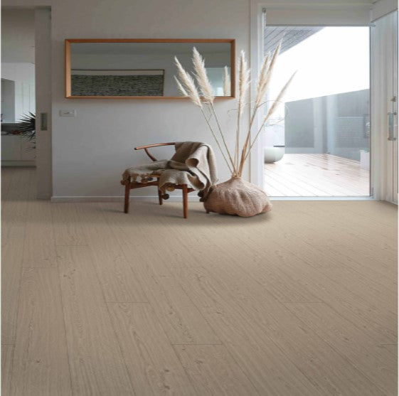 A minimalistic room with light wood flooring, a woven chair with a blanket, a large vase of pampas grass, a wall mirror, and a sliding glass door leading to a balcony. The space is bright and airy.