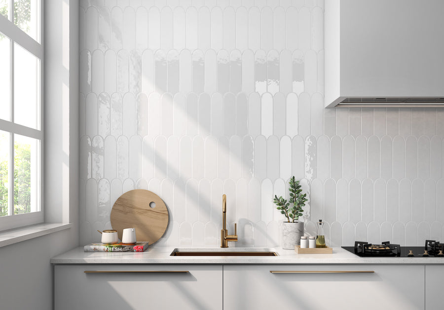 Modern white kitchen with glossy scallop tile backsplash, gold faucet, wooden cutting boards, a small plant, and a gas stove. Sunlight streams in from a large window on the left.
