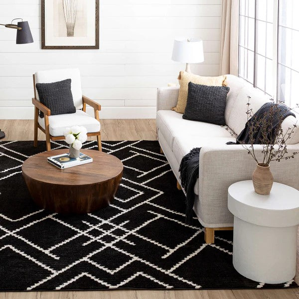 A modern living room features a beige sofa, white armchair, round wood coffee table, white side table, and large window with sheer curtains. Neutral decor is elevated by the Traverse Intersection Area Rug and contemporary accents.