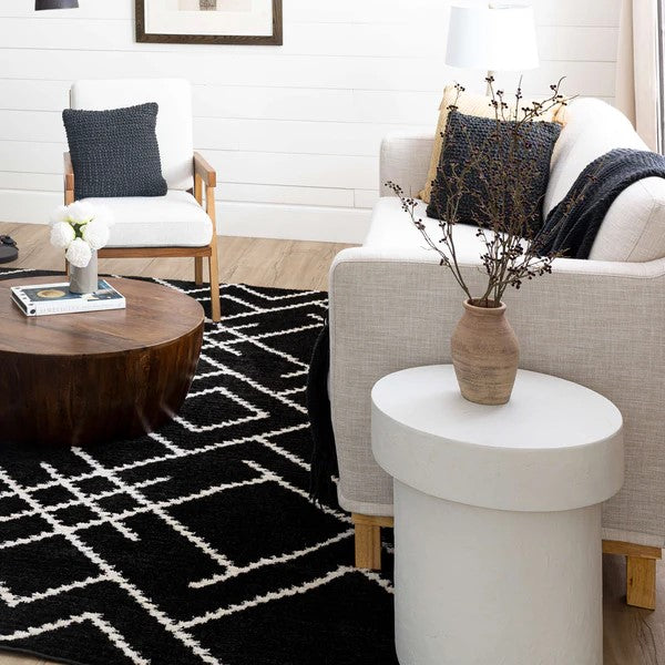 A cozy living room with a white sofa, dark pillow, wooden armchair, round wood coffee table, white side table with a vase of branches, and the Traverse Intersection Area Rug on light wood floors.