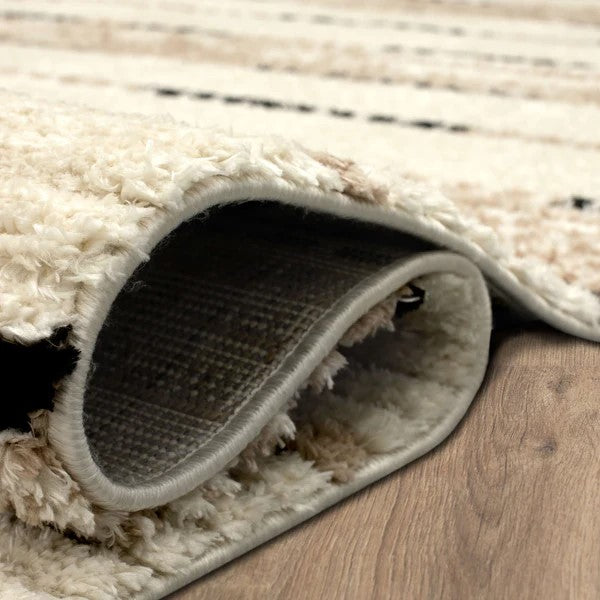 A close-up of a cream-colored, fluffy area rug with black and beige patterns, partially lifted to show its woven underside, resting on a wooden floor.