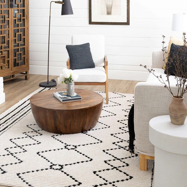 A cozy living room with a white armchair, textured pillow, round wooden coffee table, modern floor lamp, and the Traverse Paragon Area Rug featuring geometric patterns beneath décor like a vase with branches on a white side table.