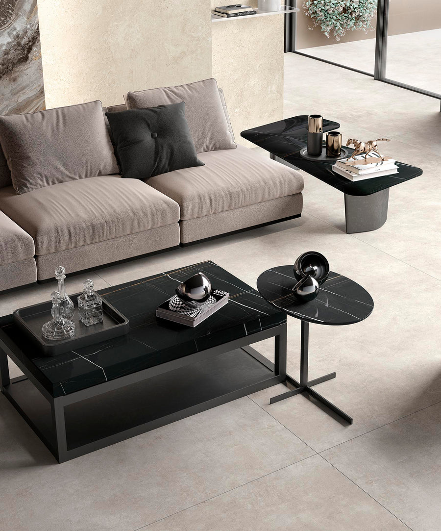 A modern living room with a beige sofa, two black marble coffee tables with metallic decor, crystal bottles, and a side table. The room features light tiled flooring and a large window with greenery outside.