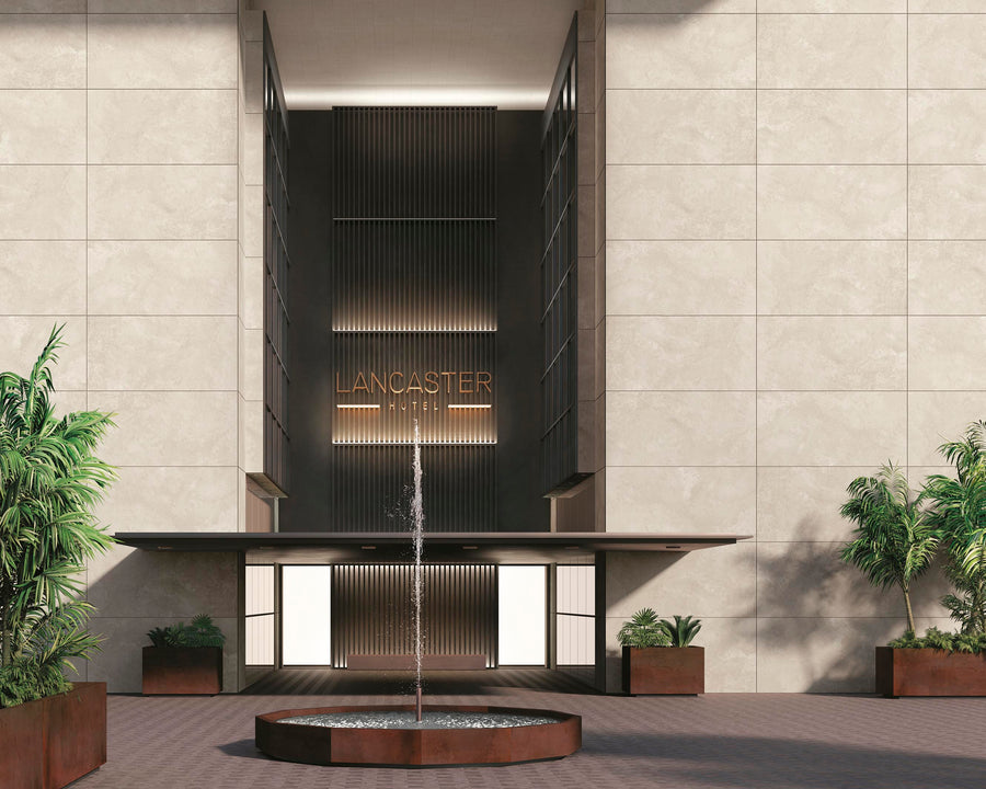 The entrance of Lancaster Hotel features a modern facade with beige walls, large windows, potted green plants, and a circular water fountain in the foreground.
