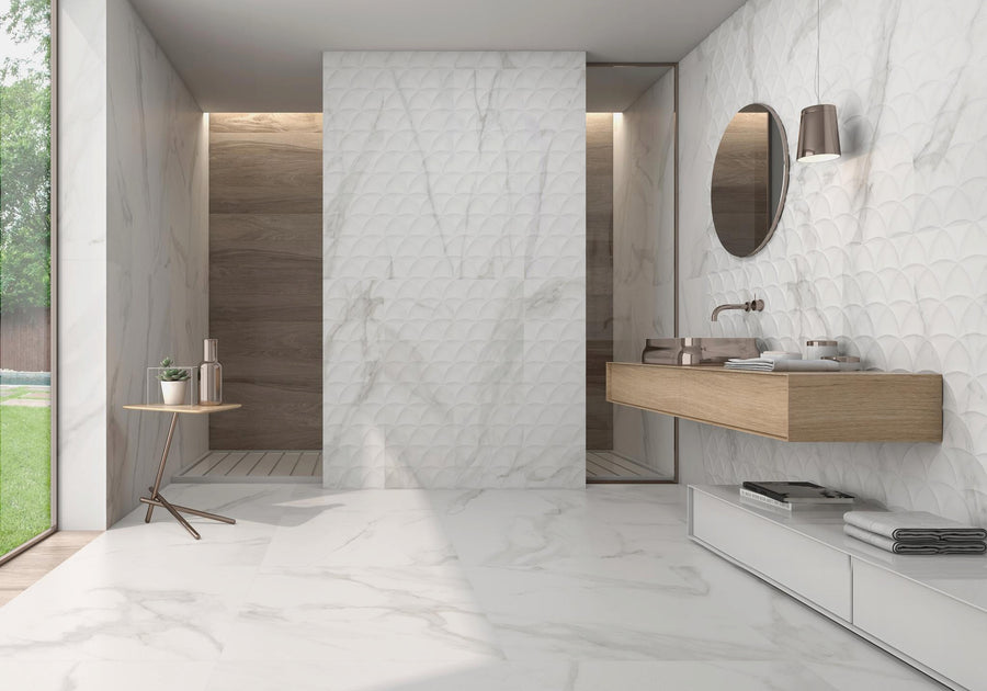 Modern bathroom with white marble walls and floor, floating wooden sink with round mirror, wall sconce, small side table with decor, and a walk-in shower area with wood accents. Large window provides natural light.