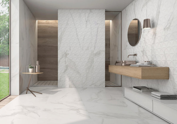 Modern bathroom with white marble walls and floor, floating wooden sink with round mirror, wall sconce, small side table with decor, and a walk-in shower area with wood accents. Large window provides natural light.