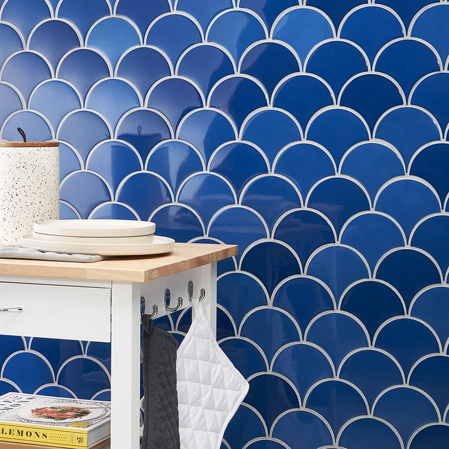 A kitchen corner with a small table, stacked plates, and a towel holder, set against a wall of glossy blue fish scale tiles with white grout.