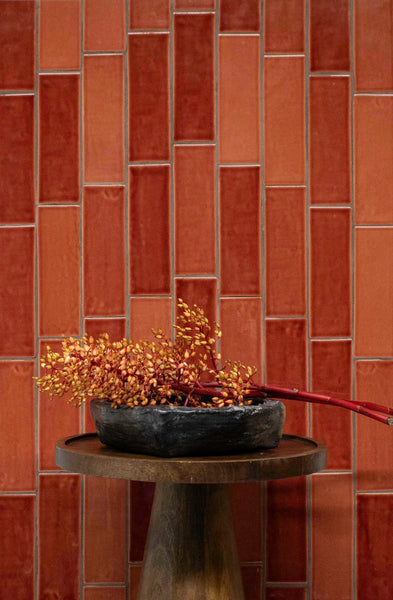 A dark bowl with orange berries sits on a round wooden table in front of a vertical red tile wall.
