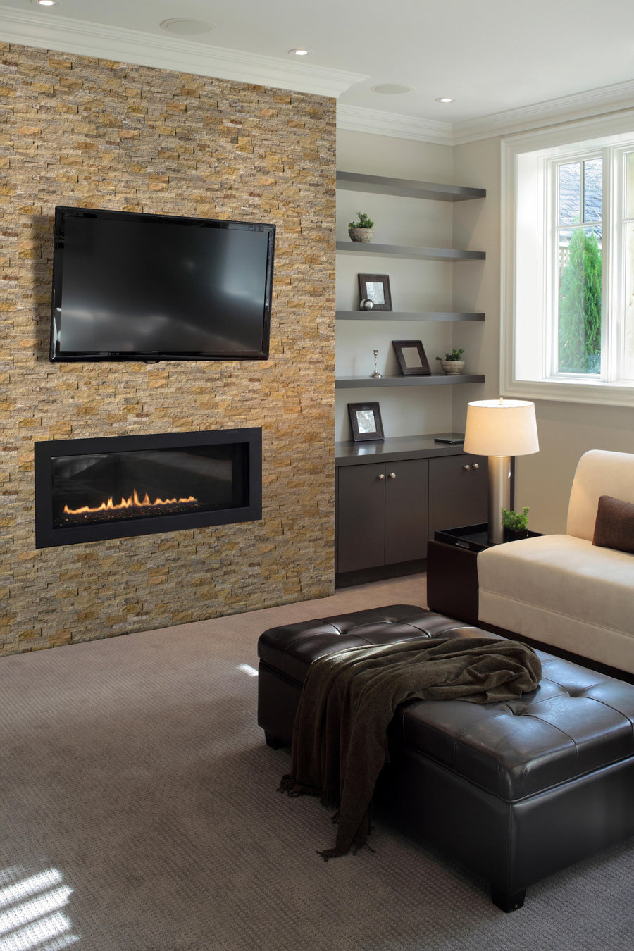 A modern living room with a stone accent wall featuring a mounted flat-screen TV and a sleek gas fireplace below. There are shelves with decor items, a sofa, a lamp, and a leather ottoman with a throw blanket.