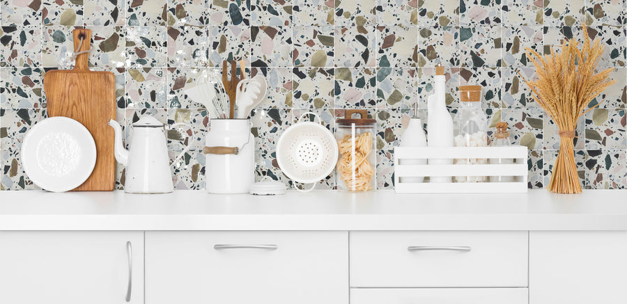 A modern white kitchen countertop with various utensils, cutting boards, jars, bottles, and a bundle of wheat, set against a terrazzo-style backsplash.
