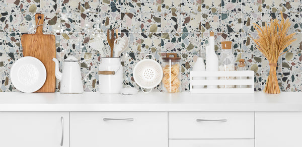 A modern white kitchen countertop with various utensils, cutting boards, jars, bottles, and a bundle of wheat, set against a terrazzo-style backsplash.